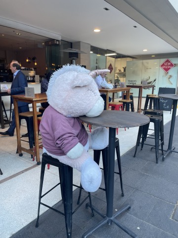 a large stuffed unicorn in a t-shirt sits alone at a tall table on the sidewalk outside a bar